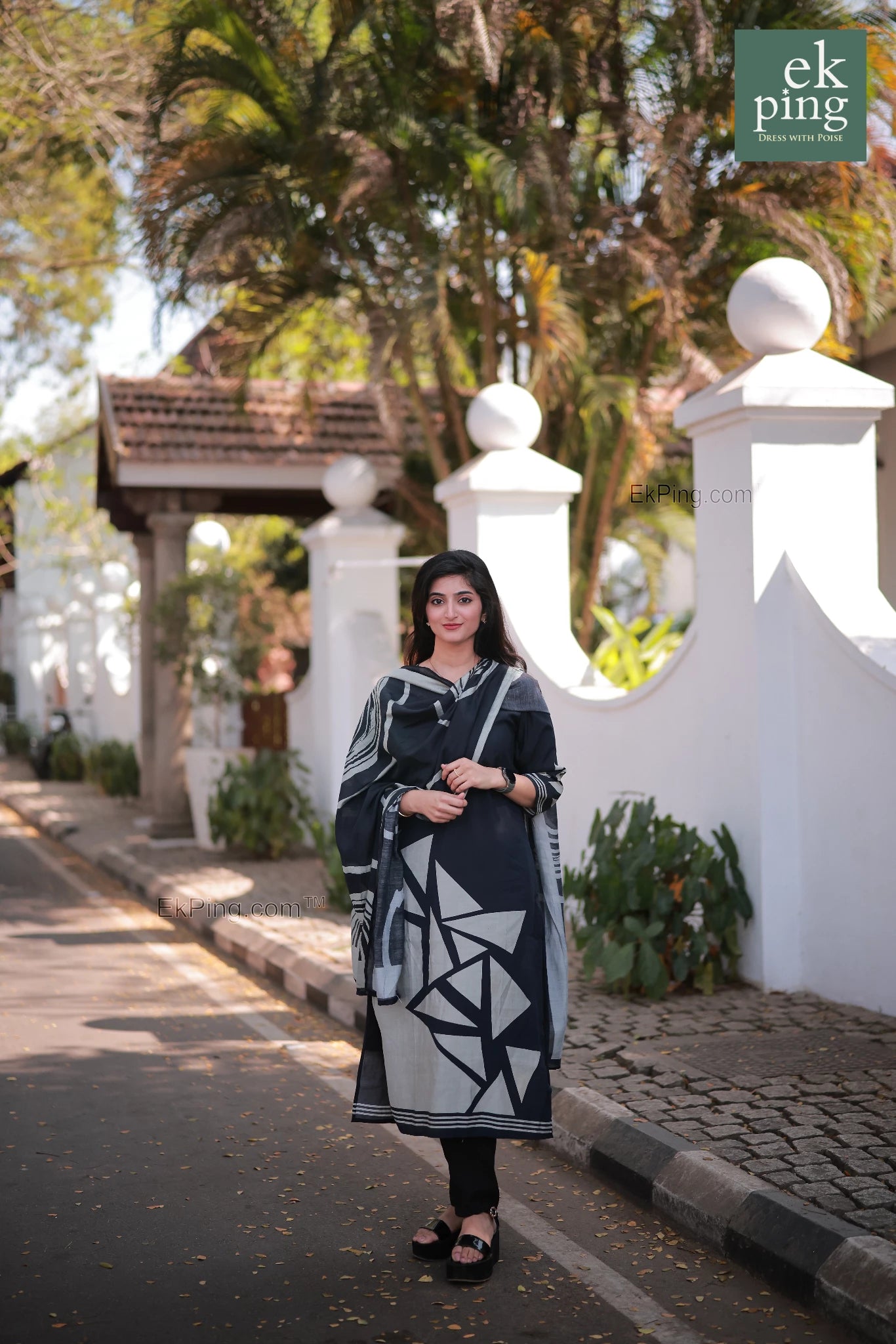 Woman in a black and white patterned office wear salwar set standing on a street with trees and a white wall in the background.