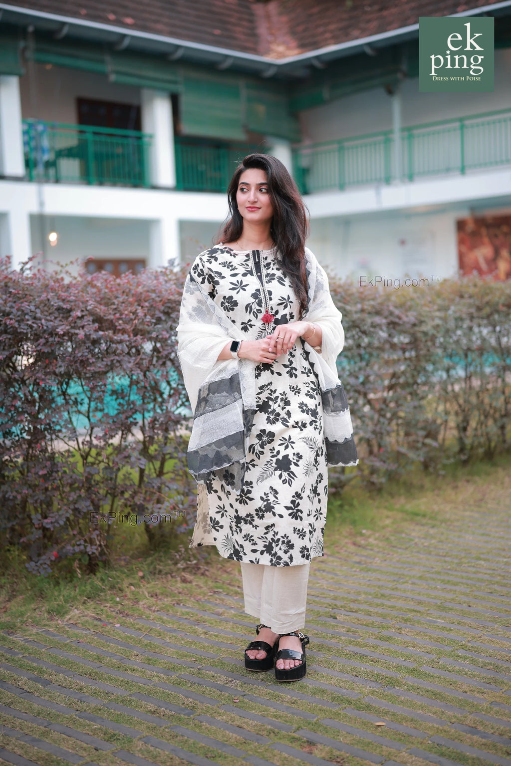 Woman in a white and black floral Salwar Set standing outdoors with a building and trees in the background