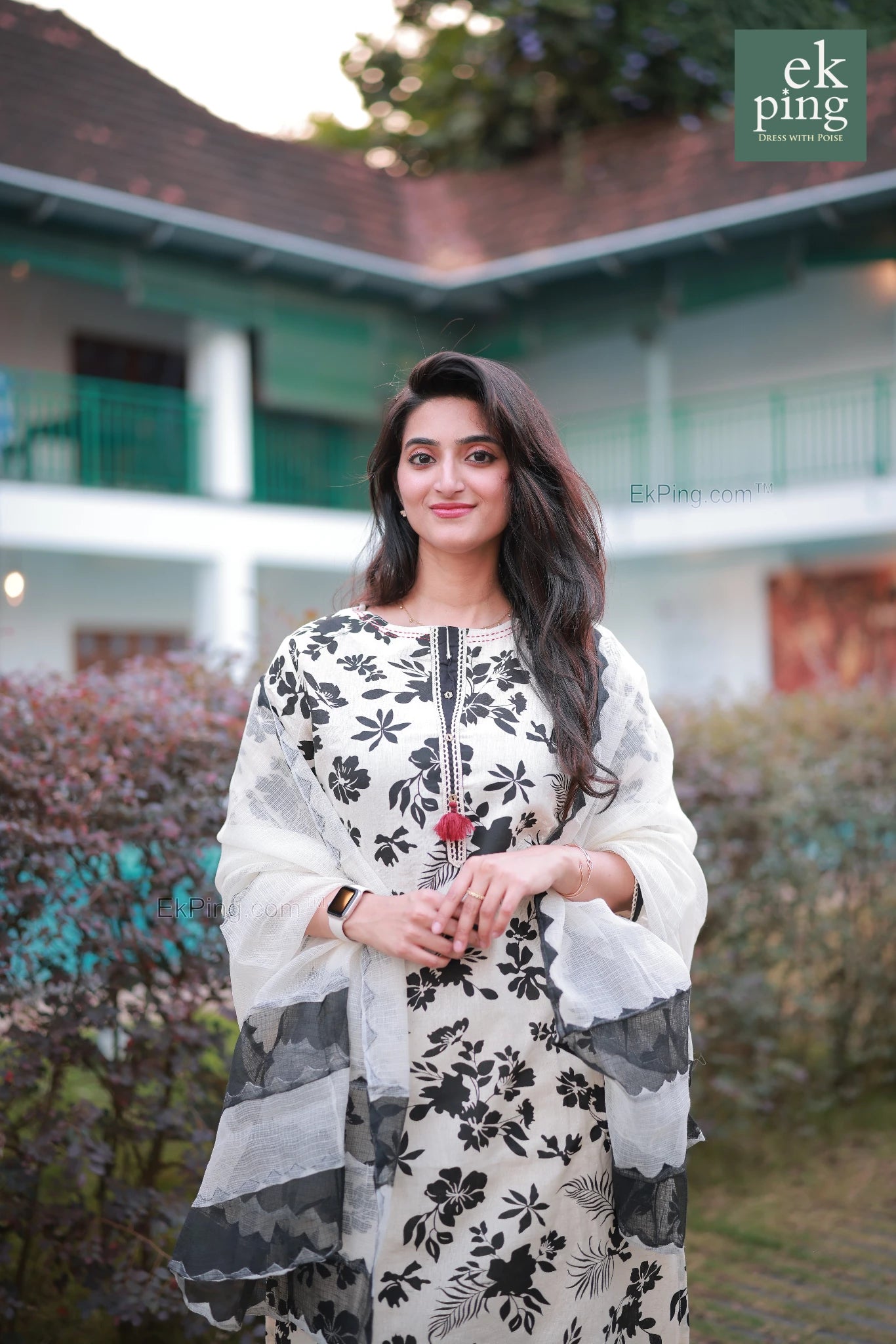 Woman in a white and black floral Salwar Set standing outdoors with a building and greenery in the background