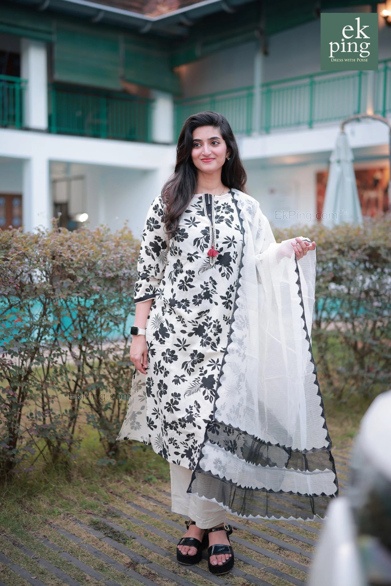 Woman in a white and black floral Salwar Set standing outdoors with a building in the background.