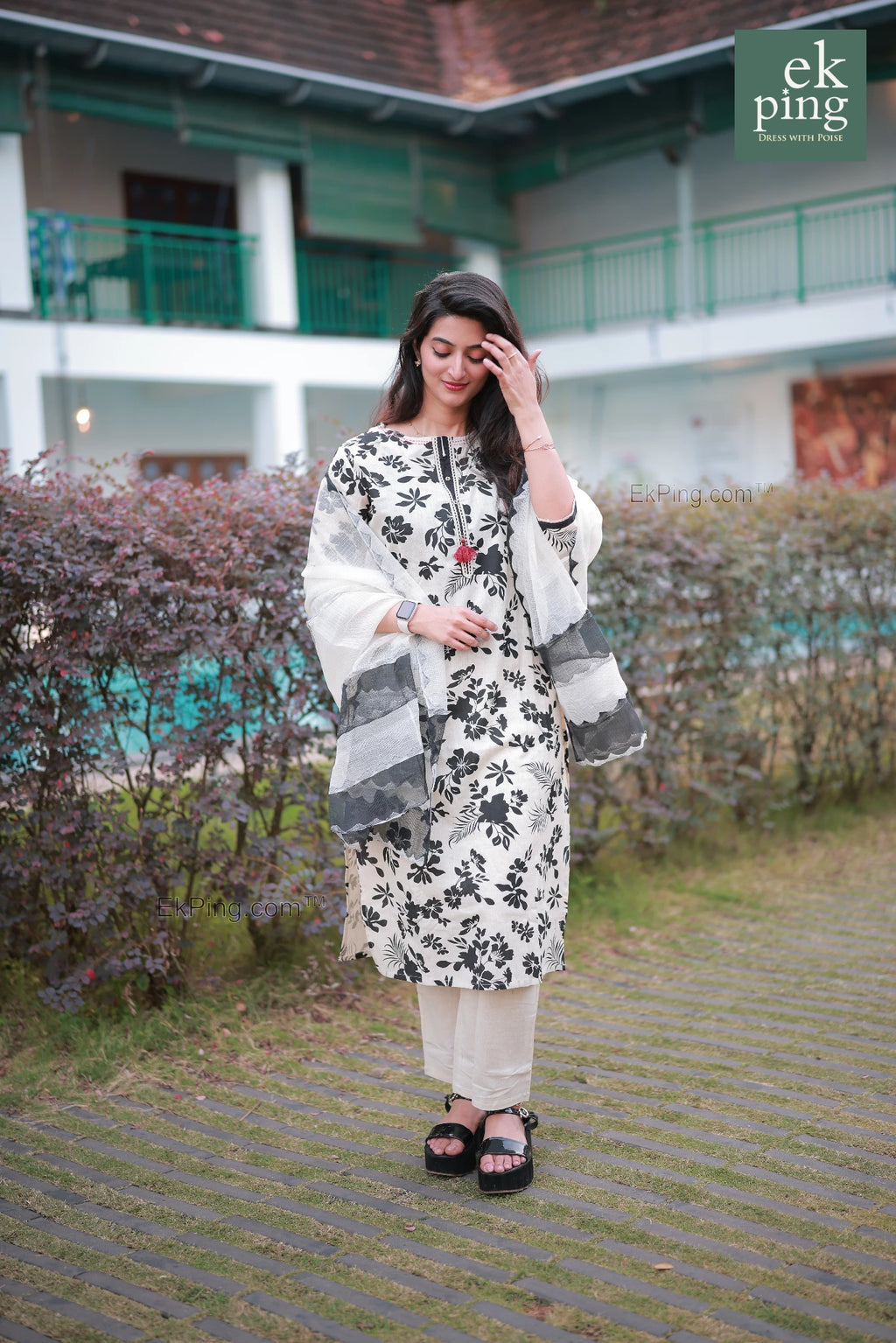 Woman in a white and black floral Salwar Set standing outdoors with a building and greenery in the background