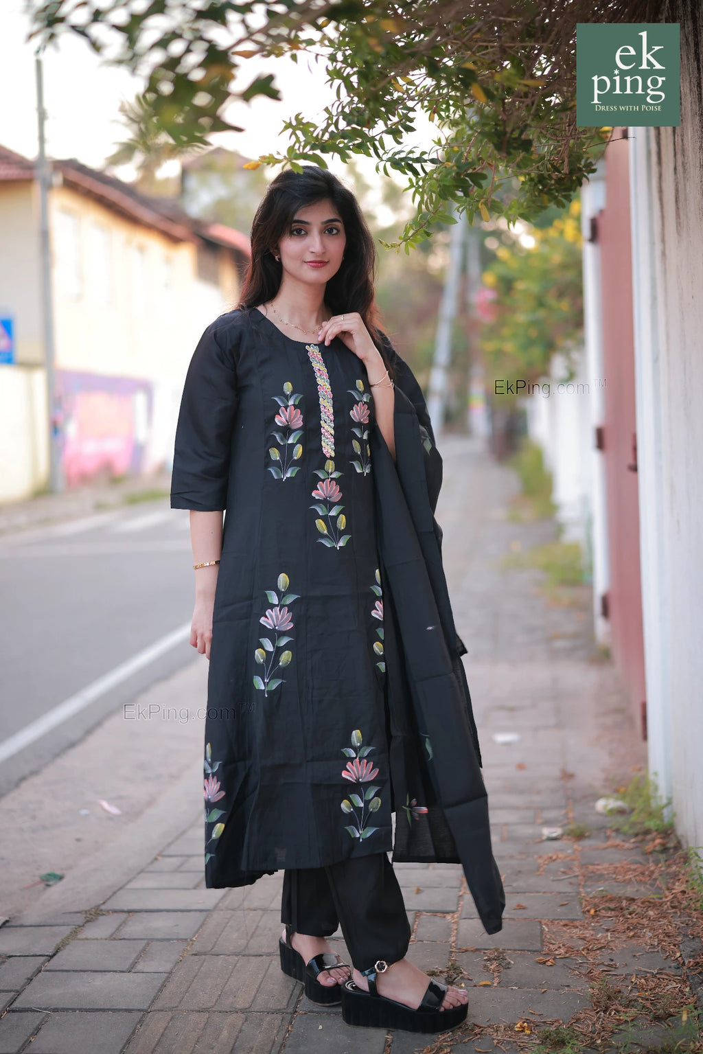 Woman wearing a black salwar set standing on a sidewalk with greenery in the background