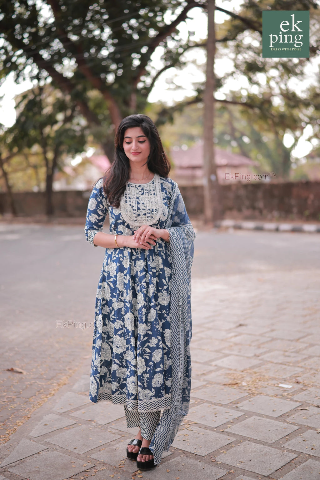 Woman in a floral anarkali dress standing on a paved road with trees in the background