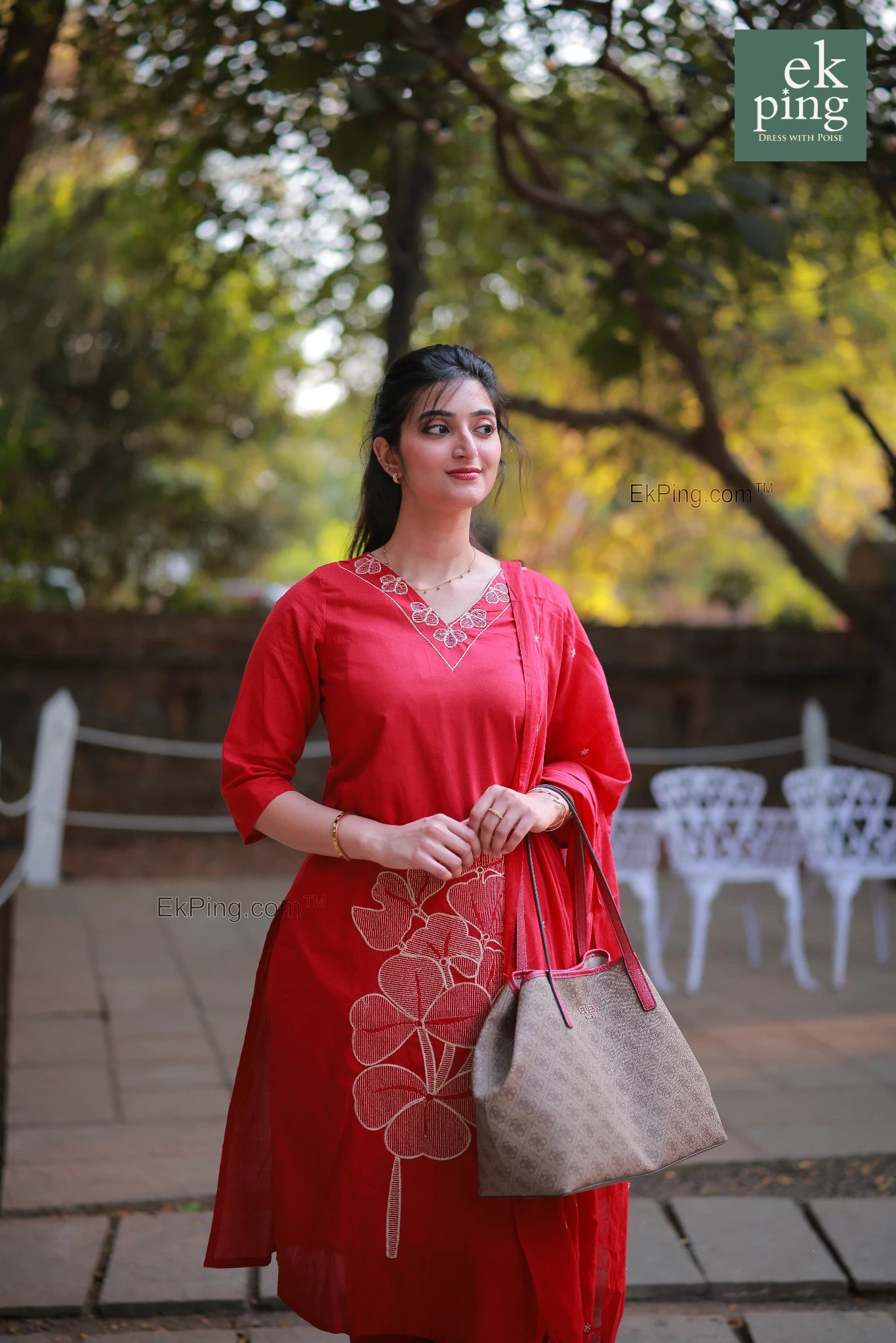 Woman in a red tkurti standing outdoors with trees and chairs in the background.