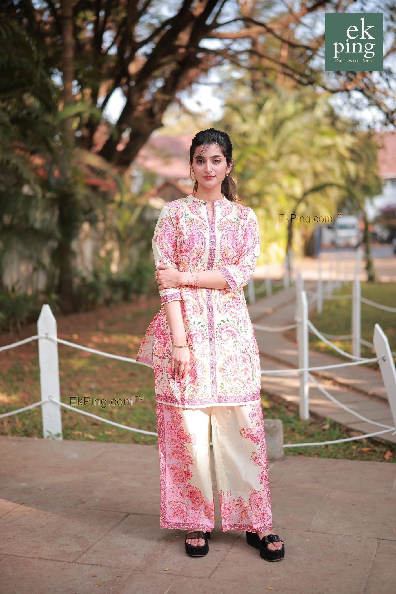 Woman in a pink floral co-ord set with palazzo pants standing outdoors with trees and a path in the background.