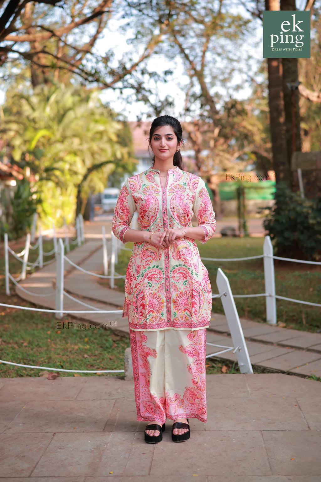 Woman in a pink floral co-ord set with palazzo pants standing outdoors with trees and a pathway in the background.