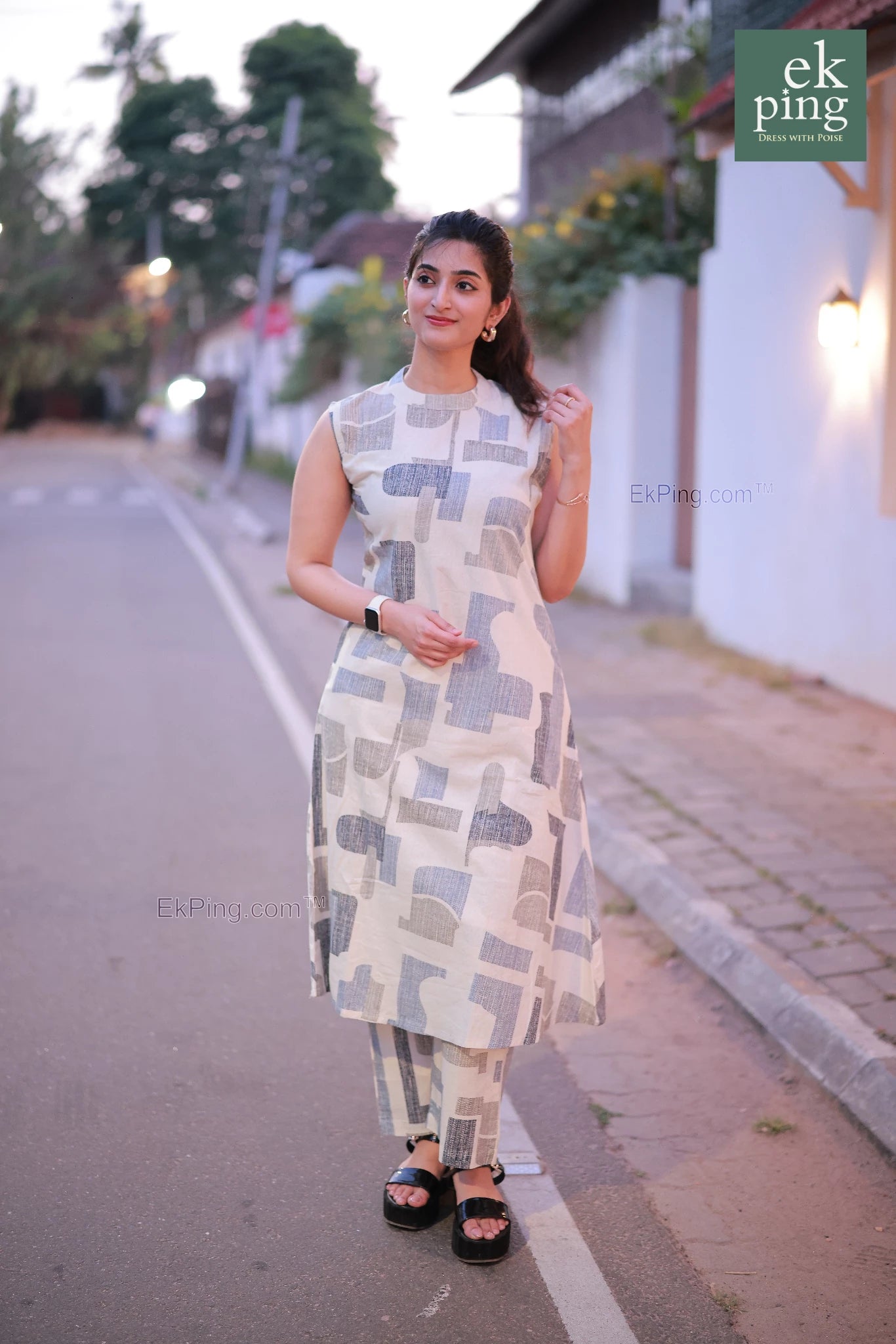 Woman in a white co-ord set dress standing on a street with a building and trees in the background.