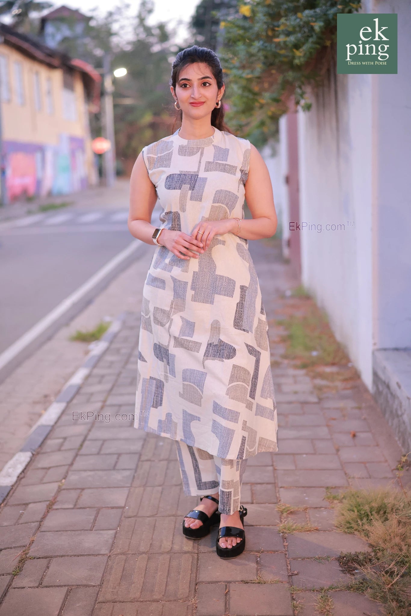 Woman in a white co-ord set dress standing on a sidewalk with a street and building in the background.