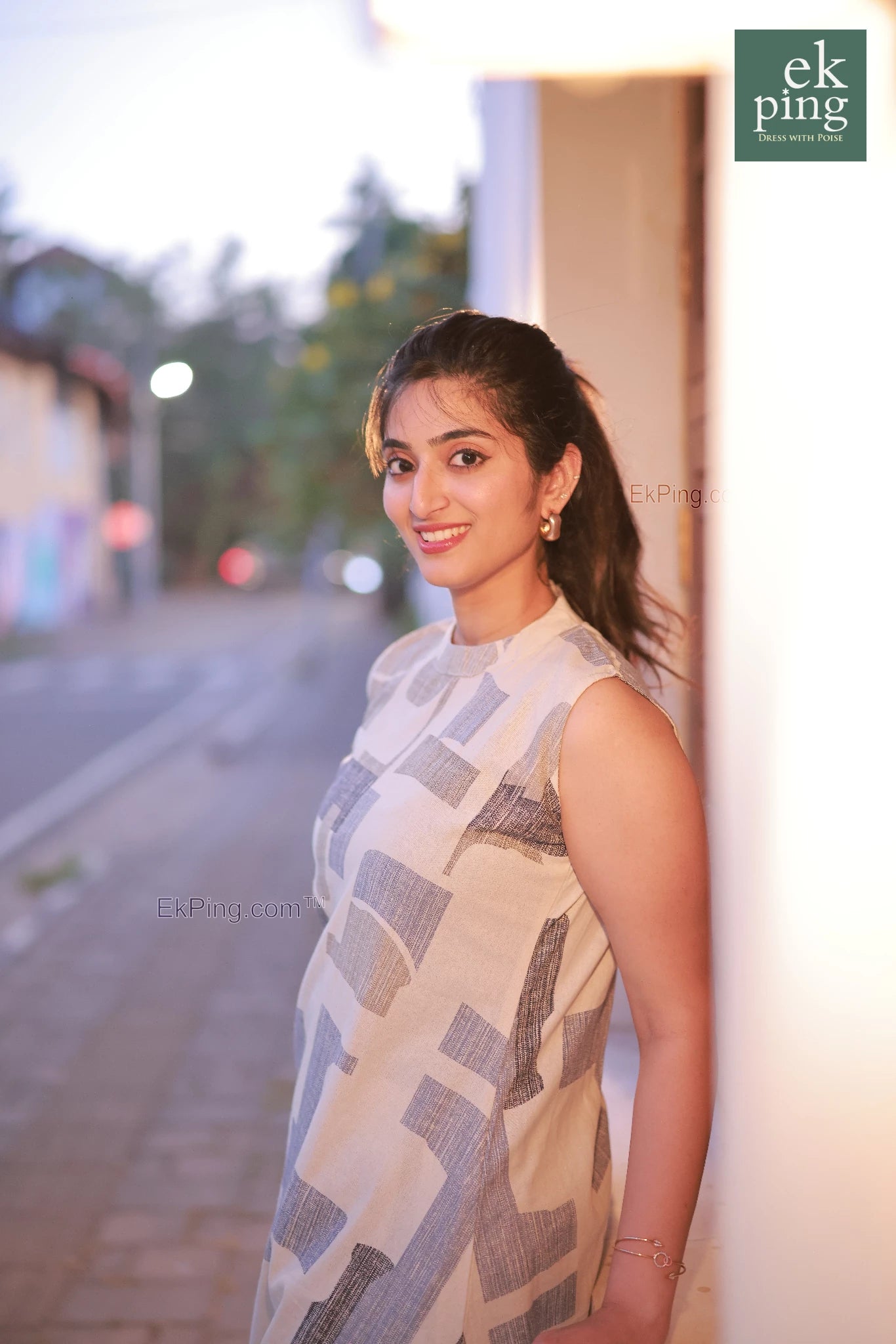 Woman in a patterned white co-ord set dress leaning against a wall with a blurred street background