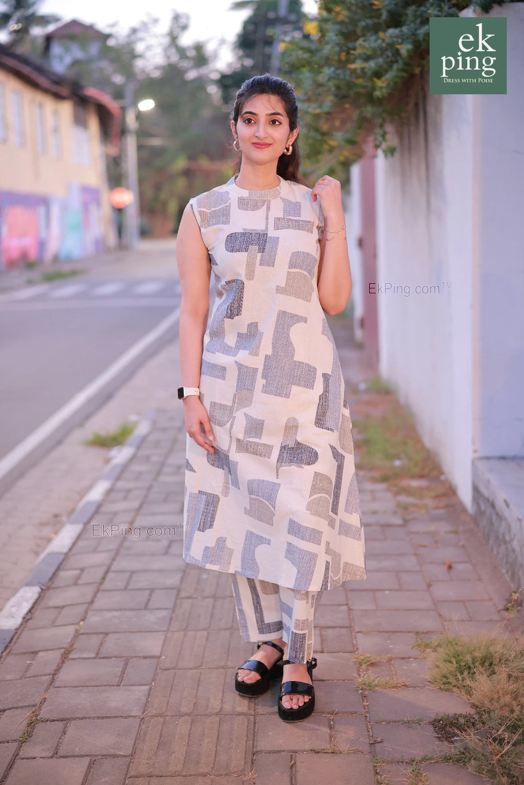 Woman in a white co-ord set dress standing on a sidewalk with a street and building in the background.