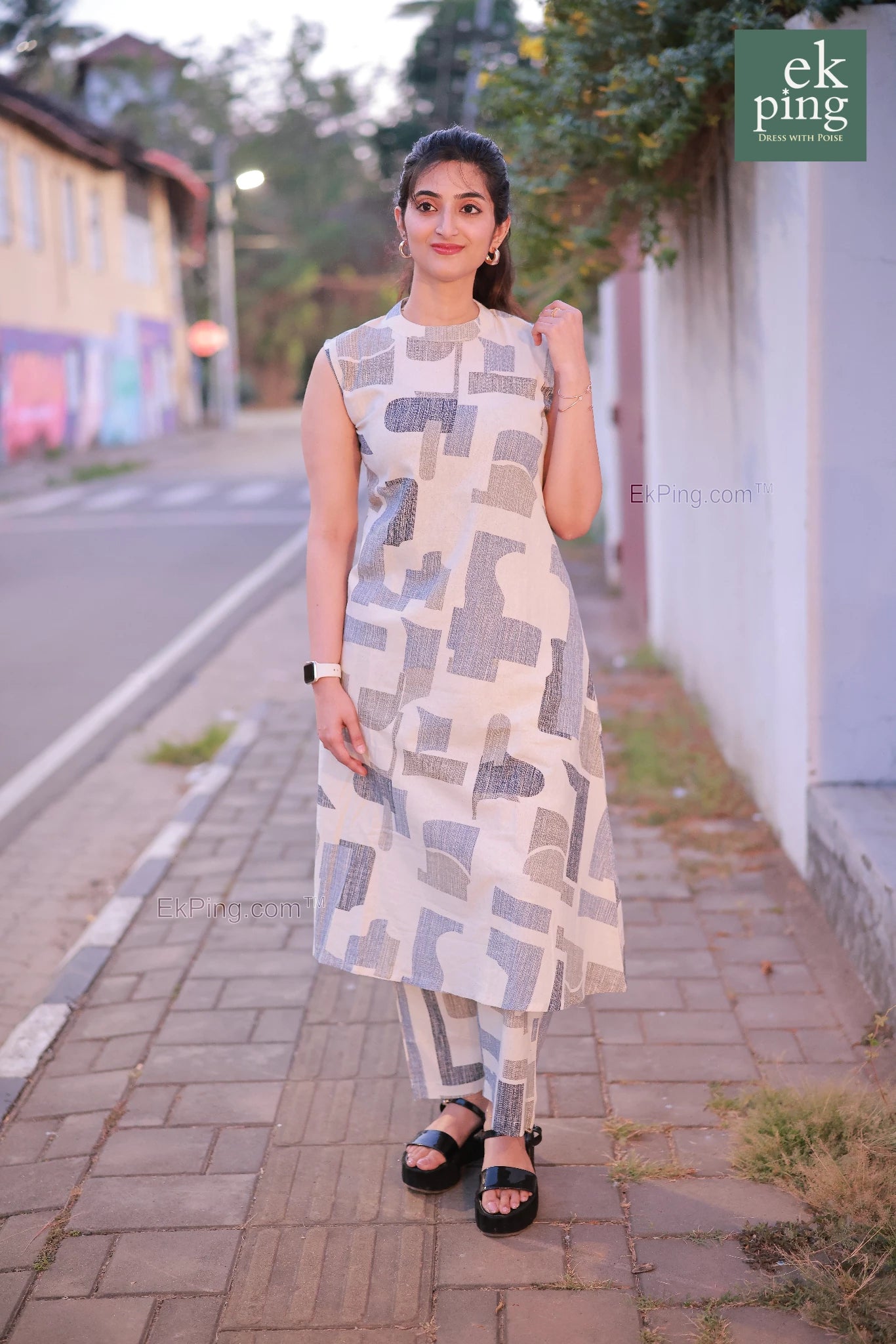 Woman in a white co-ord set dress standing on a sidewalk with a street and building in the background.