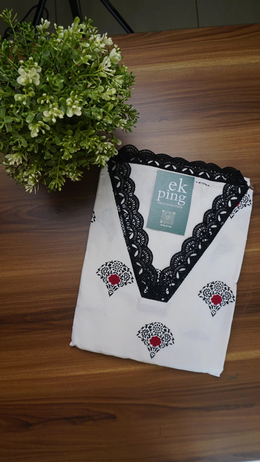 White kurti top with black lace and red floral patterns on a wooden table with flower vase nearby